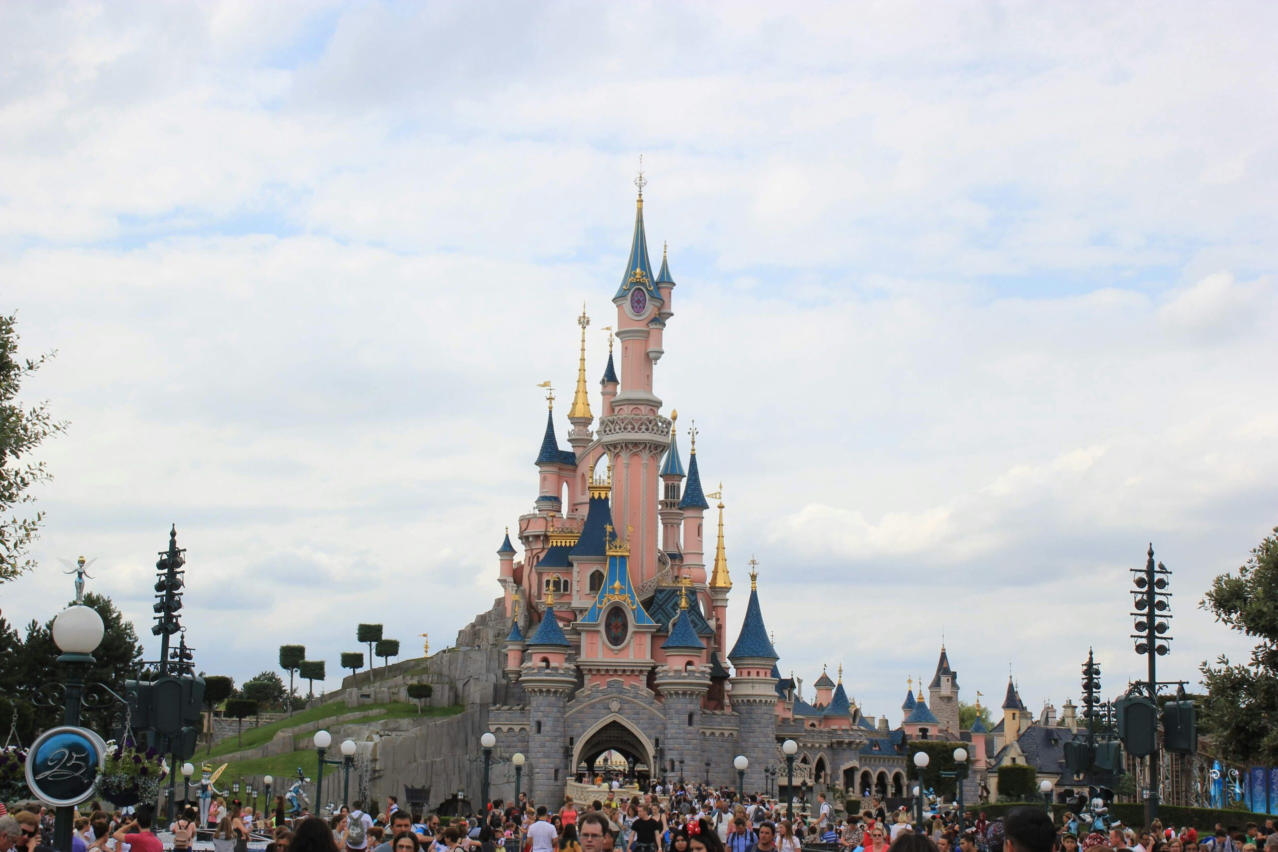 Une foule nombreuse se rassemble devant un château de conte de fées rose aux toits bleus, sous un ciel partiellement nuageux à Disneyland Paris, alors que les visiteurs empruntent les files d'attente pour profiter de la magie du parc d'attractions.