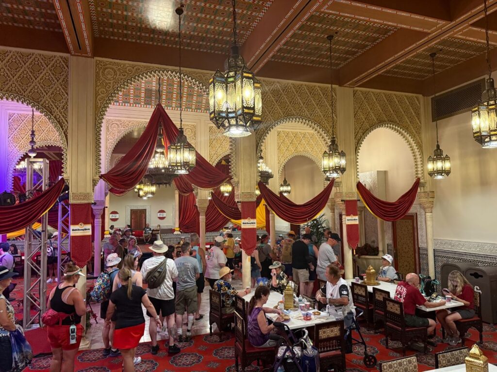Un restaurant très fréquenté dans le pavillon du Maroc d'Epcot, avec un décor orné de style moyen-oriental, des rideaux rouges, des murs à motifs, des lanternes suspendues, et des clients qui mangent ou font la queue au restaurant Marrakesh, à Disney World Orlando.