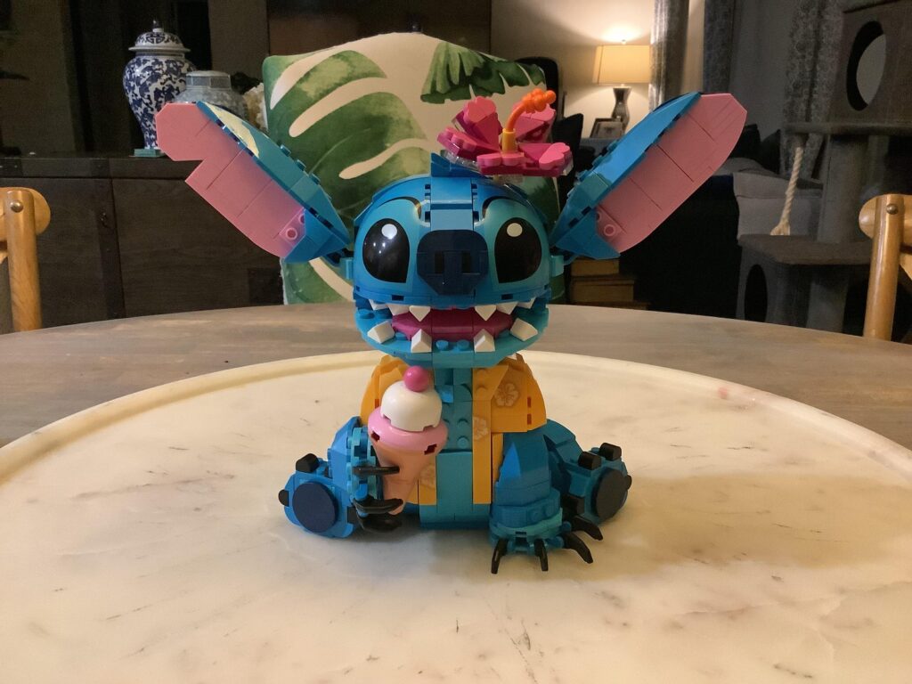 Un modèle LEGO de Stitch de Lilo & Stitch, tenant un cornet de glace et portant une fleur rose sur la tête, est présenté sur une table ronde en marbre.