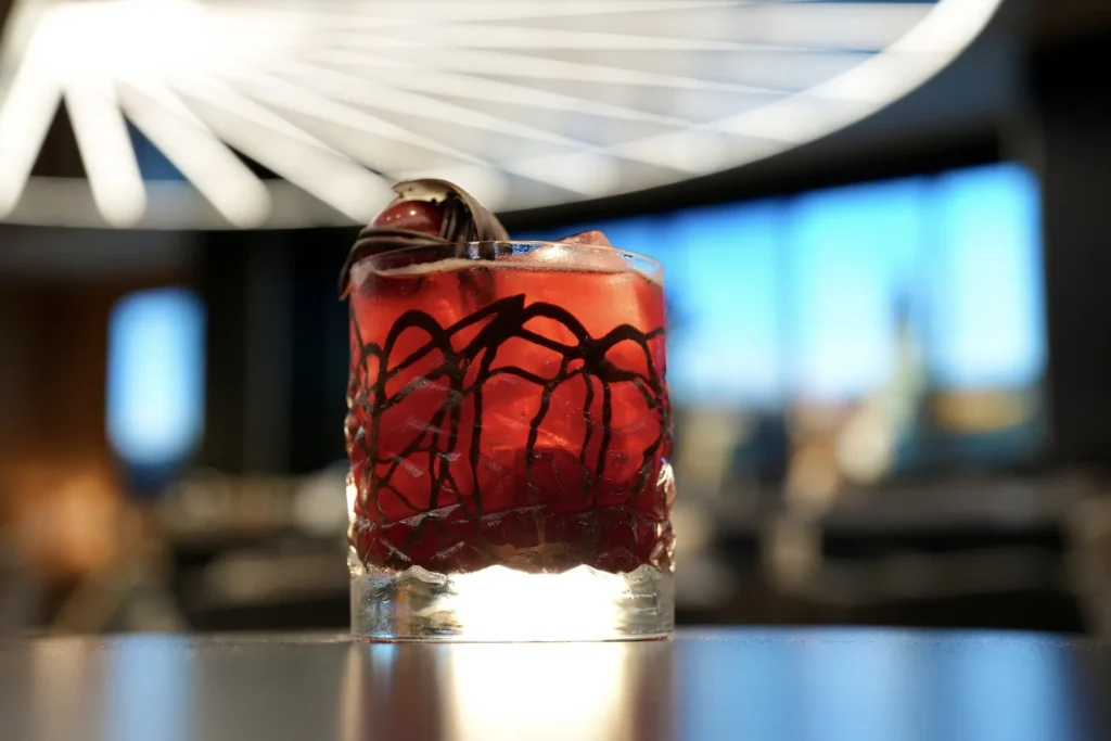 Un verre rempli d'un cocktail rouge, de glace et d'une garniture est posé sur une table à l'arrière-plan moderne et flou dans un hôtel élégant de New York, sous un luminaire géométrique.