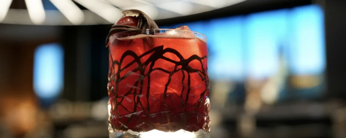 Un verre rempli d'un cocktail rouge, de glace et d'une garniture est posé sur une table à l'arrière-plan moderne et flou dans un hôtel élégant de New York, sous un luminaire géométrique.