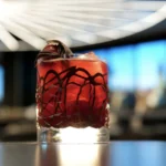 Un verre rempli d'un cocktail rouge, de glace et d'une garniture est posé sur une table à l'arrière-plan moderne et flou dans un hôtel élégant de New York, sous un luminaire géométrique.