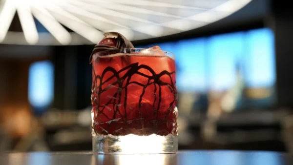 Un verre rempli d'un cocktail rouge, de glace et d'une garniture est posé sur une table à l'arrière-plan moderne et flou dans un hôtel élégant de New York, sous un luminaire géométrique.