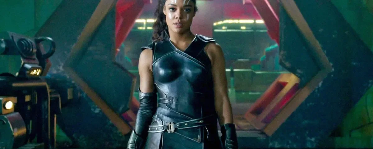 Un personnage puissant en armure noire futuriste se tient avec assurance devant une grande porte hexagonale, évoquant l'aura de Valkyrie Marvel et laissant entrevoir des origines mystérieuses dans ce décor de science-fiction.