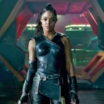 Un personnage puissant en armure noire futuriste se tient avec assurance devant une grande porte hexagonale, évoquant l'aura de Valkyrie Marvel et laissant entrevoir des origines mystérieuses dans ce décor de science-fiction.