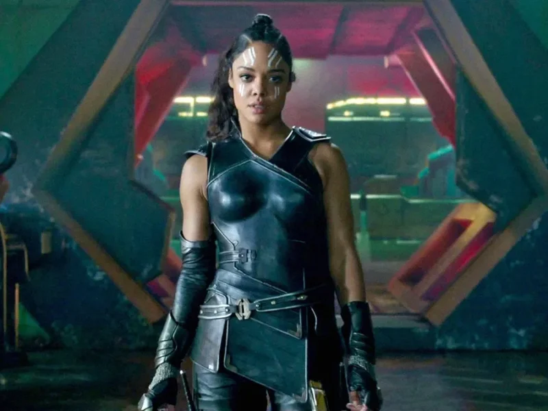 Un personnage puissant en armure noire futuriste se tient avec assurance devant une grande porte hexagonale, évoquant l'aura de Valkyrie Marvel et laissant entrevoir des origines mystérieuses dans ce décor de science-fiction.