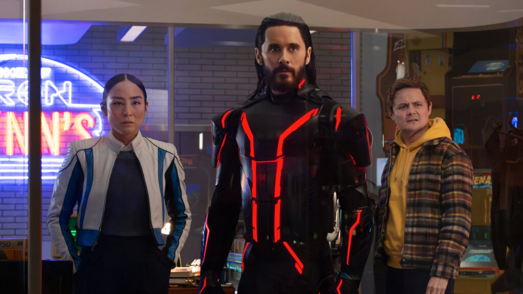 Trois personnes se tiennent à l'intérieur ; la figure centrale, qui rappelle Tron Ares, porte un costume noir avec des lignes rouges lumineuses, tandis que les deux autres semblent préoccupés. Des néons et des machines d'arcade, évoquant une franchise classique de Disney, sont visibles à l'arrière-plan.