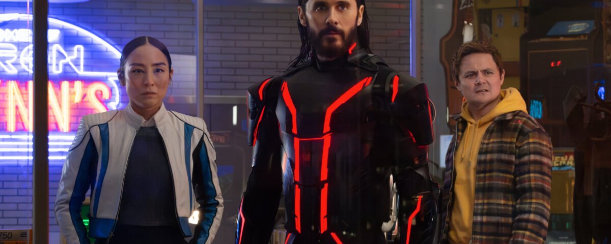 Trois personnes se tiennent à l'intérieur ; la figure centrale, qui rappelle Tron Ares, porte un costume noir avec des lignes rouges lumineuses, tandis que les deux autres semblent préoccupés. Des néons et des machines d'arcade, évoquant une franchise classique de Disney, sont visibles à l'arrière-plan.