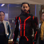 Trois personnes se tiennent à l'intérieur ; la figure centrale, qui rappelle Tron Ares, porte un costume noir avec des lignes rouges lumineuses, tandis que les deux autres semblent préoccupés. Des néons et des machines d'arcade, évoquant une franchise classique de Disney, sont visibles à l'arrière-plan.