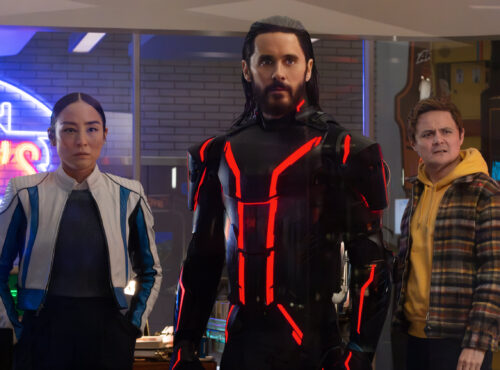 Trois personnes se tiennent à l'intérieur ; la figure centrale, qui rappelle Tron Ares, porte un costume noir avec des lignes rouges lumineuses, tandis que les deux autres semblent préoccupés. Des néons et des machines d'arcade, évoquant une franchise classique de Disney, sont visibles à l'arrière-plan.