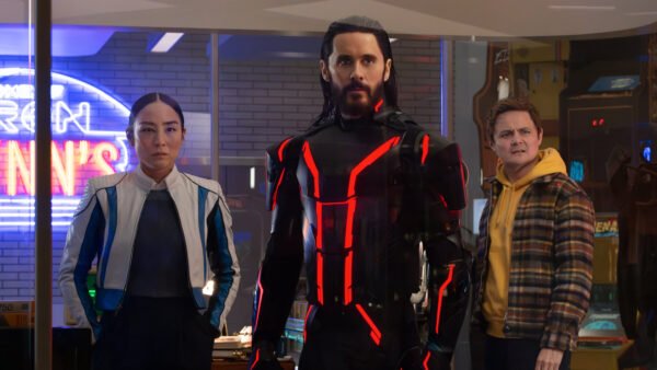 Trois personnes se tiennent à l'intérieur ; la figure centrale, qui rappelle Tron Ares, porte un costume noir avec des lignes rouges lumineuses, tandis que les deux autres semblent préoccupés. Des néons et des machines d'arcade, évoquant une franchise classique de Disney, sont visibles à l'arrière-plan.