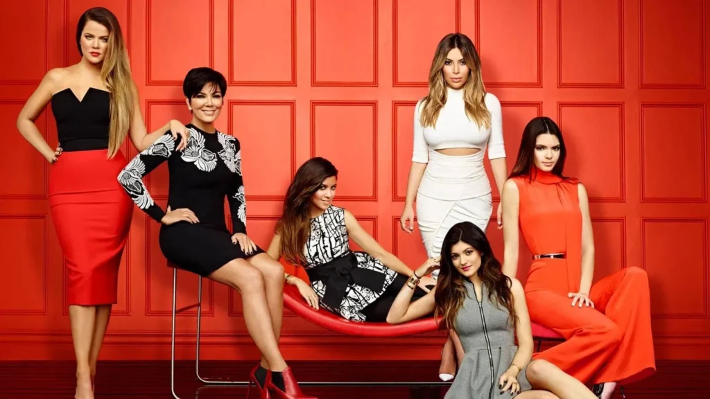 Six femmes posent ensemble devant un mur à panneaux rouges, évoquant l'ambiance bande-annonce de la Saison 7 de Kardashian, avec des tenues coordonnées rouges, noires, blanches et grises. Quatre sont debout et deux sont assises sur un banc rouge.