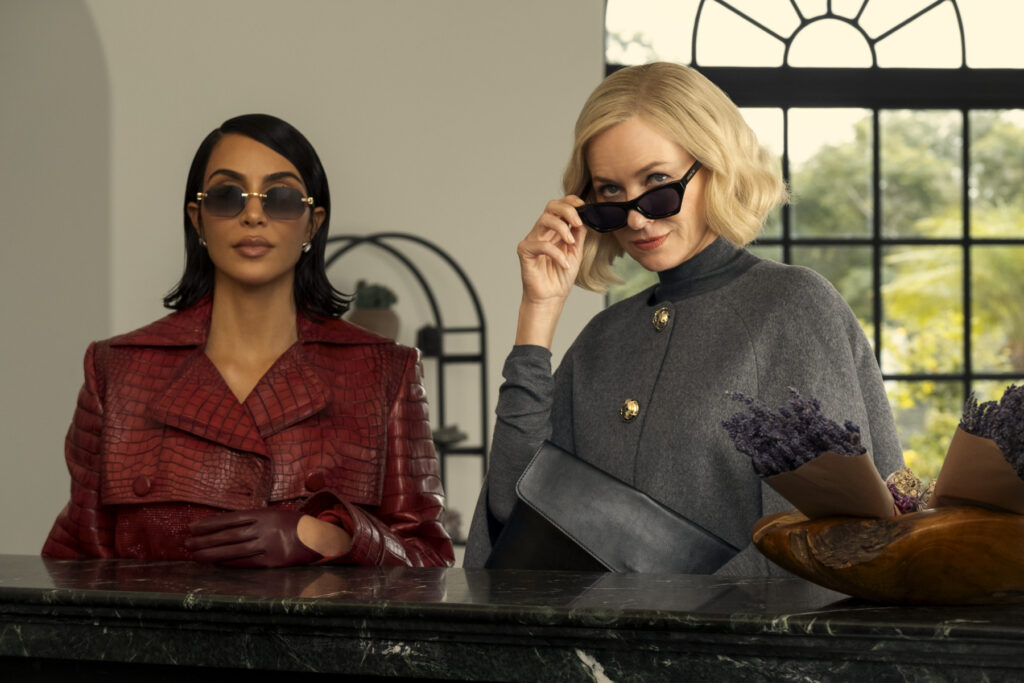 Deux femmes portant des lunettes de soleil se tiennent derrière un comptoir en marbre foncé ; l'une porte un ensemble en cuir rouge, l'autre une cape grise et une pochette noire, la lumière de la fenêtre mettant en valeur leur glamour digne de Kim Kardashian.