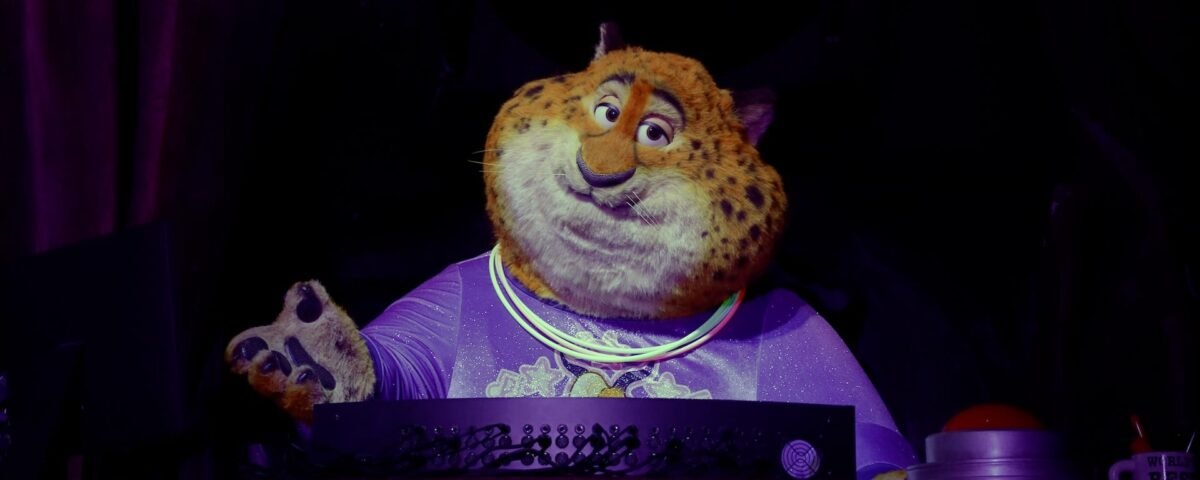 Un personnage costumé ressemblant à Clawhauser de Zootopia, portant une chemise violette et des colliers lumineux, se tient derrière une console dans un décor faiblement éclairé d'Animal Kingdom.