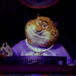 Un personnage costumé ressemblant à Clawhauser de Zootopia, portant une chemise violette et des colliers lumineux, se tient derrière une console dans un décor faiblement éclairé d'Animal Kingdom.
