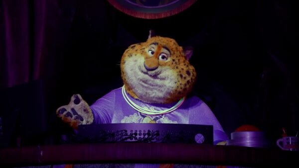 Un personnage costumé ressemblant à Clawhauser de Zootopia, portant une chemise violette et des colliers lumineux, se tient derrière une console dans un décor faiblement éclairé d'Animal Kingdom.