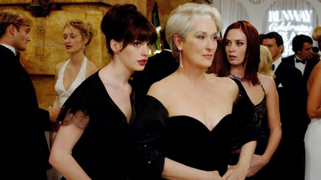 Trois femmes en tenue noire se tiennent ensemble lors d'une soirée élégante, rayonnant de la sophistication de Le Diable S'habille En Prada, avec d'autres invités se mêlant à l'arrière-plan.