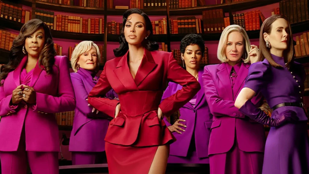 Six femmes se tiennent avec assurance devant des étagères, l'une d'entre elles en tailleur rouge au centre et les autres en tailleur violet assorti à ses côtés. Il s'agit d'une scène de responsabilisation tirée de la prochaine série Disney+ de Kim Kardashian, All's Fair.
