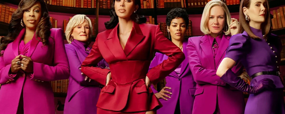 Six femmes se tiennent avec assurance devant des étagères, l'une d'entre elles en tailleur rouge au centre et les autres en tailleur violet assorti à ses côtés. Il s'agit d'une scène de responsabilisation tirée de la prochaine série Disney+ de Kim Kardashian, All's Fair.