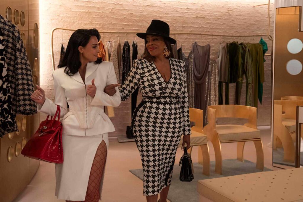 Deux femmes vêtues de tenues à la mode marchent bras dessus bras dessous dans une boutique, entourées d'étagères de vêtements et de chaises, ce qui rappelle une scène de la série glam de Disney+ "All's Fair".