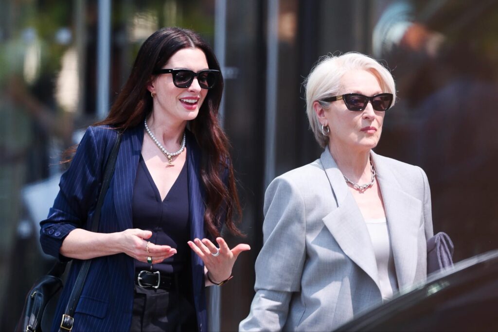 Dans le style Le Diable S'habille en Prada, deux femmes portant des lunettes de soleil et des vêtements d'affaires marchent côte à côte à l'extérieur ; l'une parle tandis que l'autre regarde devant elle.