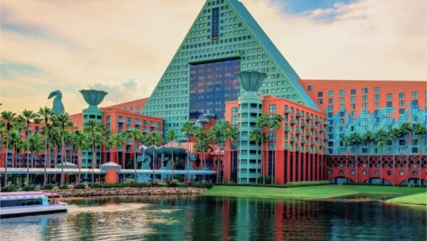 Un grand hôtel Disney World coloré avec un toit en forme de pyramide et des palmiers, situé au bord de l'eau sous un ciel nuageux, parfait pour les clients qui recherchent des forfaits spéciaux près des parcs Walt Disney.