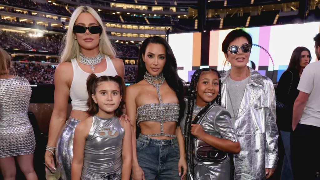 Cinq personnes se tiennent ensemble lors d'un événement. Trois adultes et deux enfants portent des tenues métalliques ou étincelantes, comme dans une scène de Kardashian Saison 7. En arrière-plan, on aperçoit la foule d'un stade.