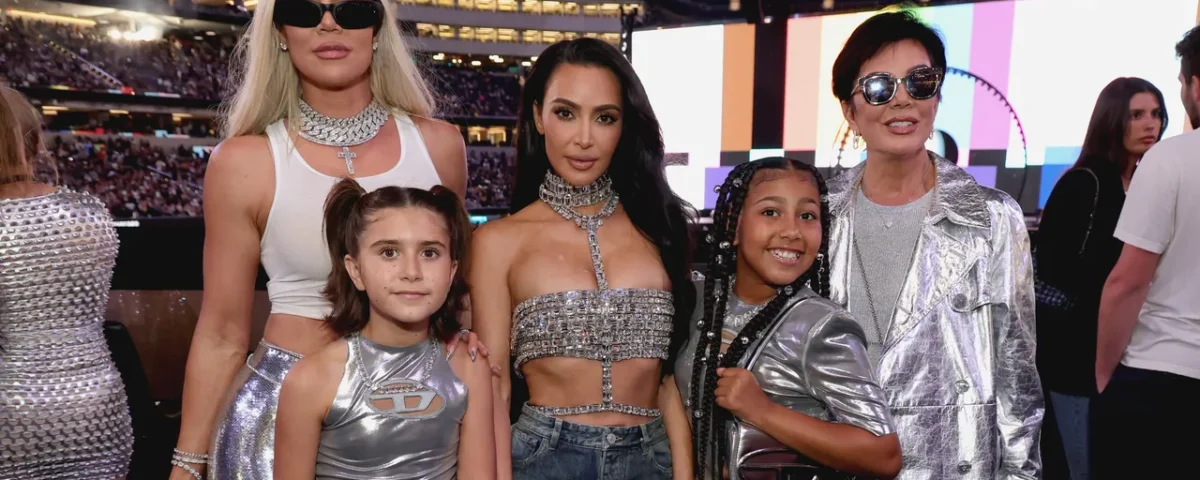 Cinq personnes se tiennent ensemble lors d'un événement. Trois adultes et deux enfants portent des tenues métalliques ou étincelantes, comme dans une scène de Kardashian Saison 7. En arrière-plan, on aperçoit la foule d'un stade.