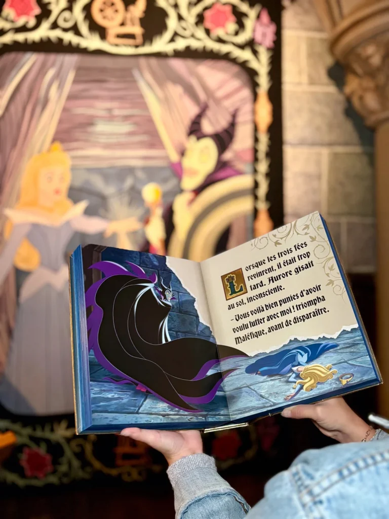 Une personne ouvre Le Grimoire De La Belle Au Bois Dormant, un livre d'histoire en Édition Limitée de Hachette Jeunesse Disney ; l'arrière-plan montre une peinture murale de la princesse Aurore de "La Belle au Bois Dormant".