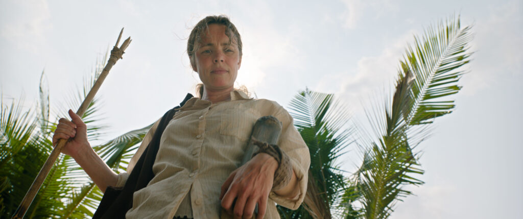 Une femme tenant une lance de fortune se tient en plein air devant des palmiers, regardant vers le bas avec une expression sérieuse, évoquant l'atmosphère tendue, huis clos d'horreur d'un thriller de Sam Raimi ou des 20th Century Studios.