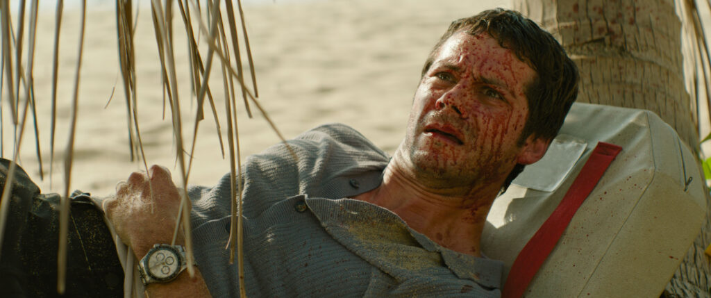 Un homme au visage et aux vêtements ensanglantés est allongé sur le sable près d'un arbre, levant les yeux avec une expression de détresse. Il porte une montre et est partiellement recouvert de sable, évoquant l'intense huis clos d'horreur souvent vu dans les thrillers des studios du XXe siècle.