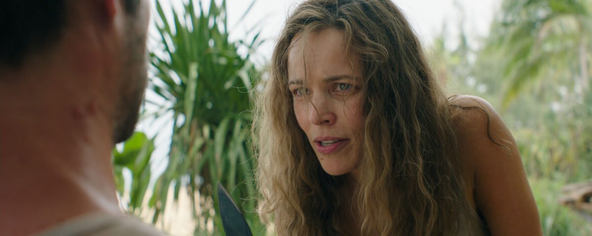 Une femme aux longs cheveux ondulés se penche en avant et regarde attentivement un homme, évoquant un huis clos d'horreur, avec des plantes vertes en arrière-plan.