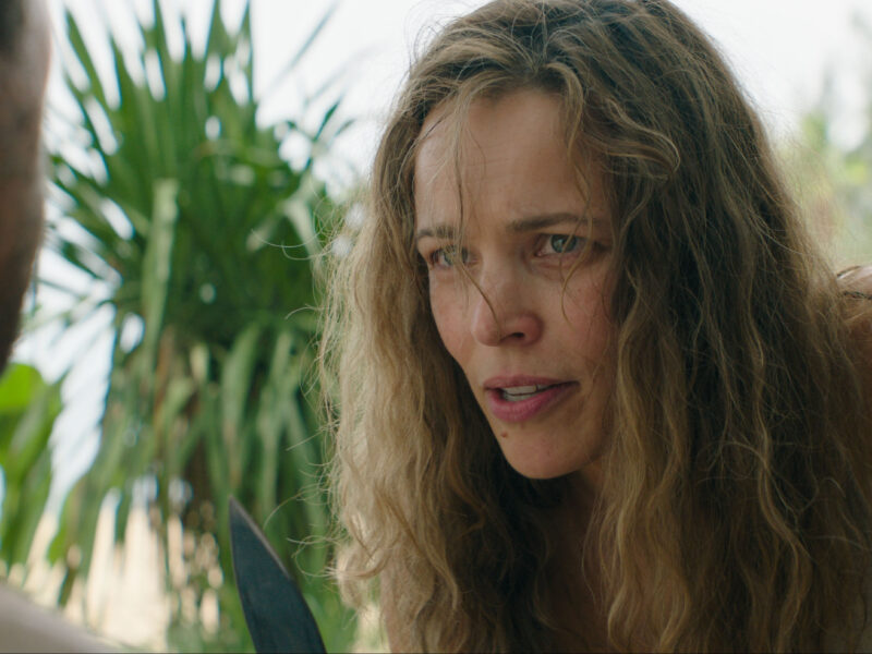 Une femme aux longs cheveux ondulés se penche en avant et regarde attentivement un homme, évoquant un huis clos d'horreur, avec des plantes vertes en arrière-plan.