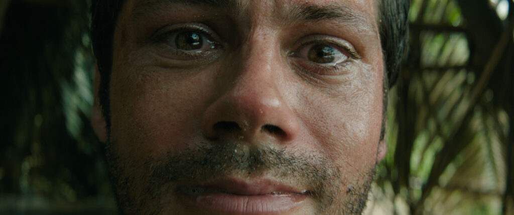 Gros plan d'un homme à la barbe hirsute et au visage mouillé, regardant attentivement devant lui - une image qui rappelle le style huis clos d'horreur de Sam Raimi ; l'arrière-plan montre un feuillage vert flou.