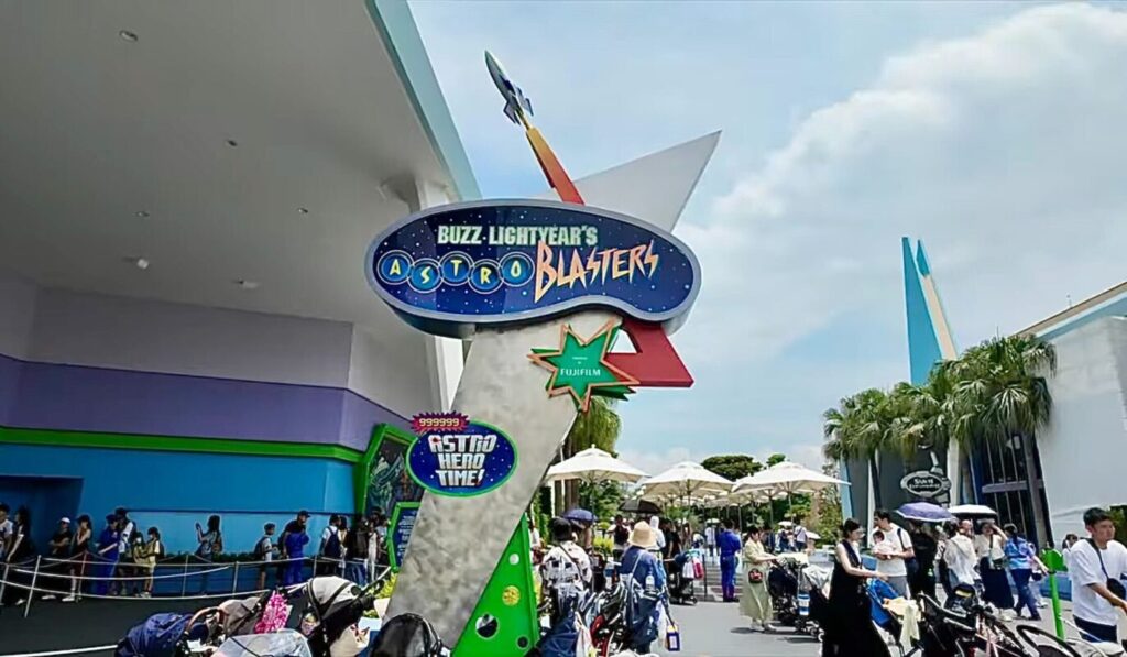 Panneau d'entrée de Buzz Lightyear's Astro Blasters à Tokyo Disneyland, avec des personnes faisant la queue et des poussettes garées à proximité dans le parc d'attractions.