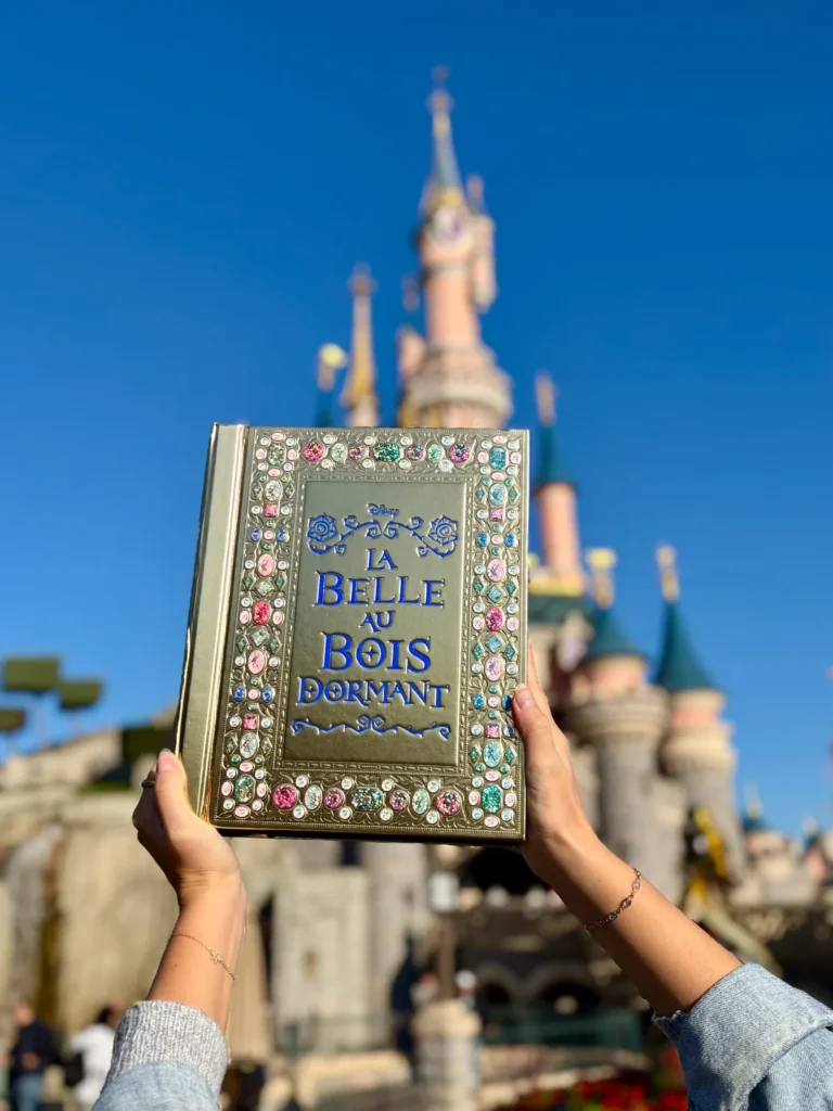 Une personne brandit Le Grimoire De La Belle Au Bois Dormant, une Édition Limitée de Hachette Jeunesse Disney, devant un château de conte de fées rose sous un ciel bleu clair.