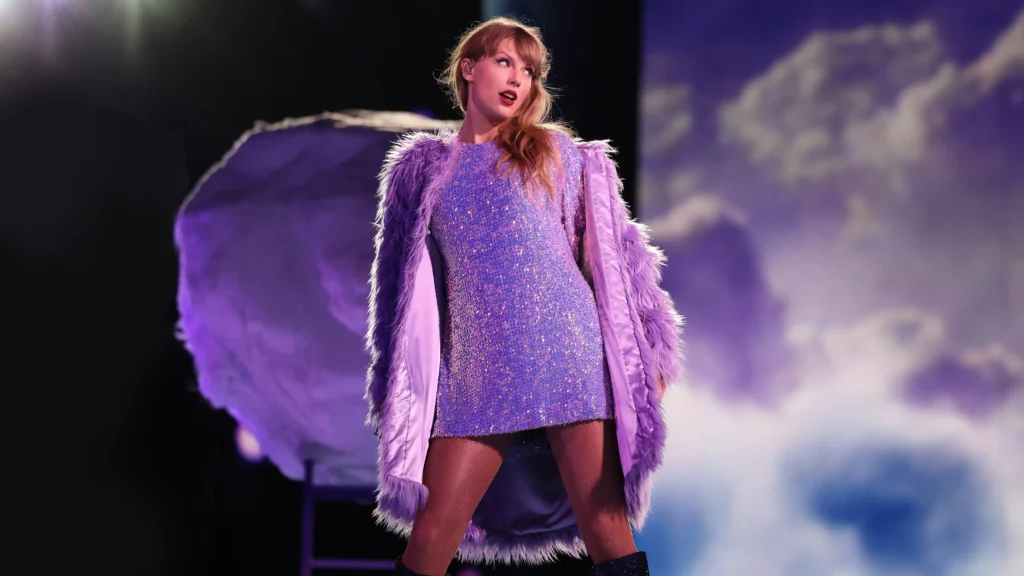 Une femme se tient sur scène, vêtue d'une robe violette étincelante et d'un manteau à plumes lavande, avec un éclairage dramatique et un arrière-plan en forme de nuage derrière elle, créant une scène qui rappelle un film-concert de Taylor Swift sur Disney+.