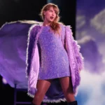 Une femme se tient sur scène, vêtue d'une robe violette étincelante et d'un manteau à plumes lavande, avec un éclairage dramatique et un arrière-plan en forme de nuage derrière elle, créant une scène qui rappelle un film-concert de Taylor Swift sur Disney+.