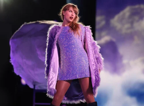 Une femme se tient sur scène, vêtue d'une robe violette étincelante et d'un manteau à plumes lavande, avec un éclairage dramatique et un arrière-plan en forme de nuage derrière elle, créant une scène qui rappelle un film-concert de Taylor Swift sur Disney+.