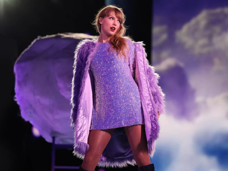 Une femme se tient sur scène, vêtue d'une robe violette étincelante et d'un manteau à plumes lavande, avec un éclairage dramatique et un arrière-plan en forme de nuage derrière elle, créant une scène qui rappelle un film-concert de Taylor Swift sur Disney+.