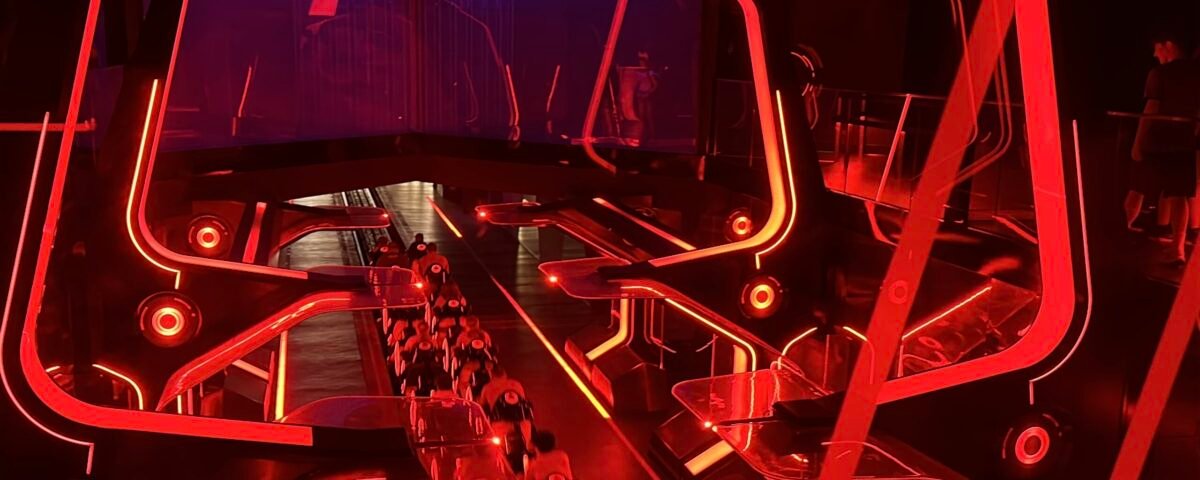 Une aire de lancement de montagnes russes intérieures futuristes s'illumine de néons rouge vif, avec des structures angulaires et des véhicules de type moto sur une piste inspirée du Lightcycle Run de Tron.