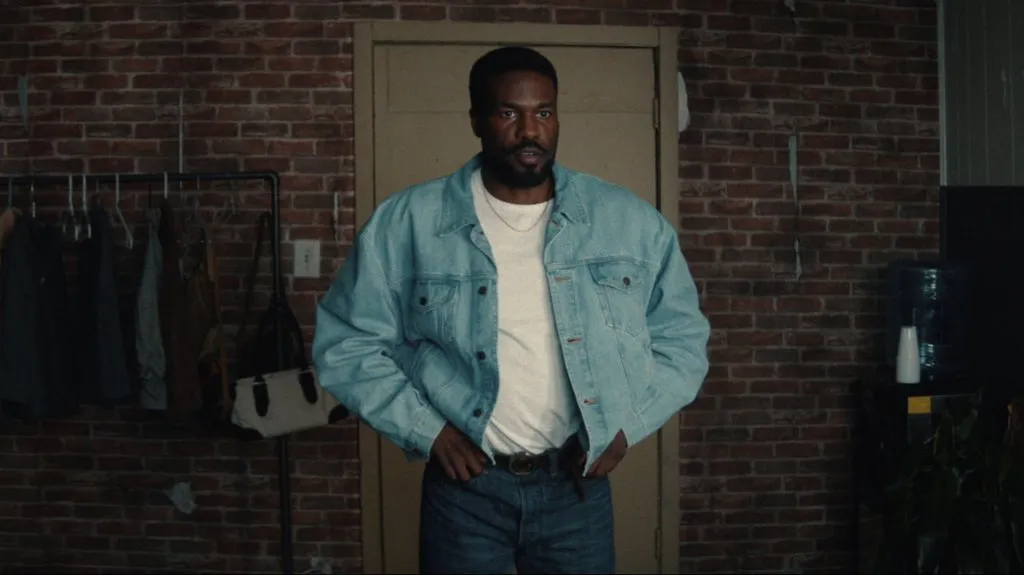 Un homme portant une veste en jean clair, un t-shirt blanc et un jean se tient à l'intérieur devant une porte fermée et un mur de briques. Il incarne l'assurance froide de Wonder Man de Marvel dans la prochaine série Disney+.