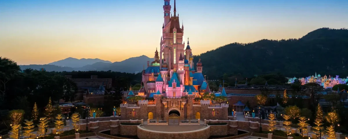 Le château de conte de fées de Hong Kong Disneyland, illuminé, se dresse au centre d'une place paysagée au crépuscule, avec les montagnes en silhouette à l'arrière-plan, offrant des aperçus magiques des attractions enchanteresses du parc.