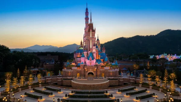 Le château de conte de fées de Hong Kong Disneyland, illuminé, se dresse au centre d'une place paysagée au crépuscule, avec les montagnes en silhouette à l'arrière-plan, offrant des aperçus magiques des attractions enchanteresses du parc.