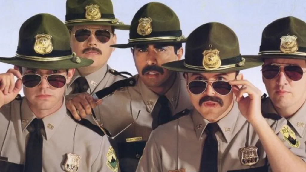 Cinq policiers en uniforme portant des chapeaux et des lunettes de soleil se tiennent près les uns des autres, face à la caméra, avec des expressions sérieuses - rappelant les Soldats de l'hiver ou une scène de Super Troopers 3.