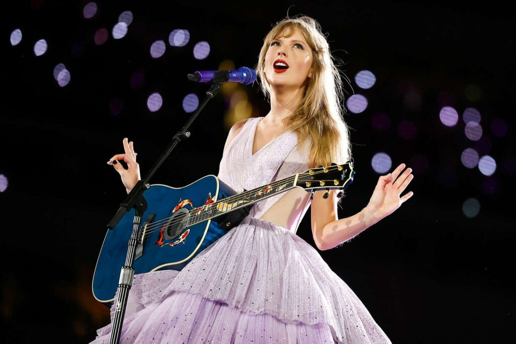 Une femme vêtue d'une robe lavande chante sur scène, jouant d'une guitare acoustique bleue sous les lumières de la scène, capturant ainsi une ambiance magique de ciné-concert, parfaite pour tout fan de Disney+ ou de Taylor Swift.