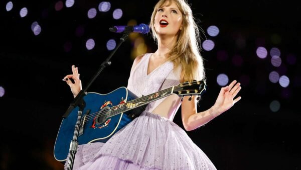 Une femme vêtue d'une robe lavande chante sur scène, jouant d'une guitare acoustique bleue sous les lumières de la scène, capturant ainsi une ambiance magique de ciné-concert, parfaite pour tout fan de Disney+ ou de Taylor Swift.