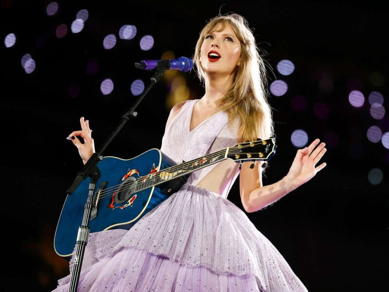 Une femme vêtue d'une robe lavande chante sur scène, jouant d'une guitare acoustique bleue sous les lumières de la scène, capturant ainsi une ambiance magique de ciné-concert, parfaite pour tout fan de Disney+ ou de Taylor Swift.
