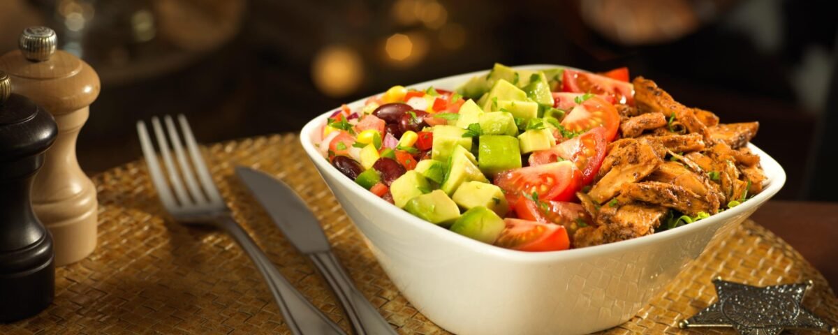 Un bol de salade avec du poulet grillé, de l'avocat, des tomates, des haricots, du maïs et de la laitue est posé sur un napperon tissé à côté d'une fourchette, d'un couteau et d'une cuillère - parfait pour ceux qui recherchent des menus végétariens comme à Disneyland Paris.