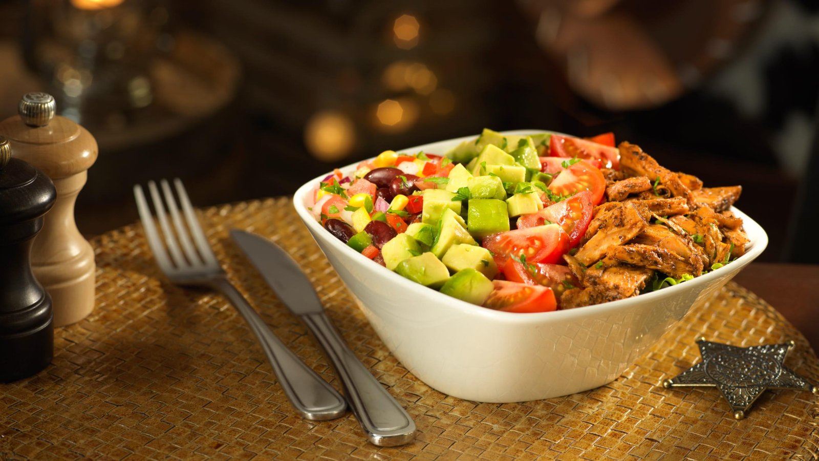 Un bol de salade avec du poulet grillé, de l'avocat, des tomates, des haricots, du maïs et de la laitue est posé sur un napperon tissé à côté d'une fourchette, d'un couteau et d'une cuillère - parfait pour ceux qui recherchent des menus végétariens comme à Disneyland Paris.
